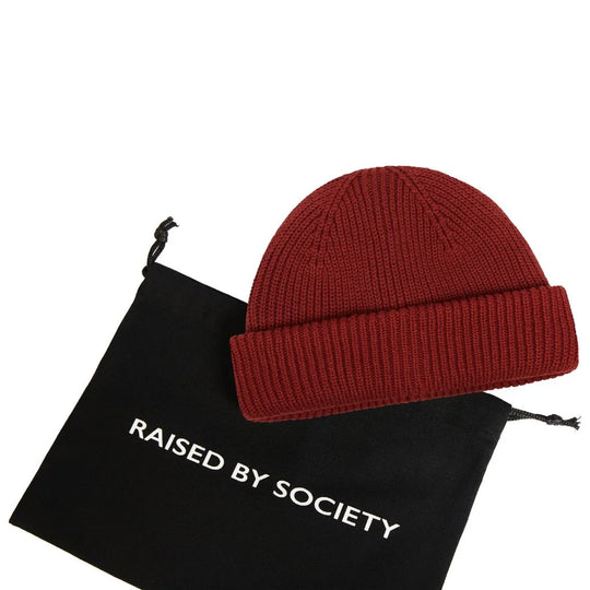 modern fisherman beanie style. Red beanie on a black drawstring bag with 'Raised by Society' branding.